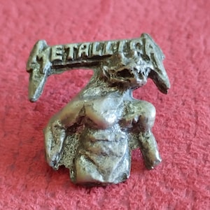 Può includere: Una spilla in metallo color argento raffigurante una figura stilizzata con la scritta "METALLICA" in alto. La figura presenta un design dettagliato e strutturato su sfondo rosso.