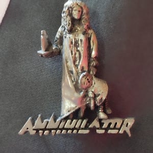 Op de afbeelding: Een zilverkleurige metalen pin met een figuur die een kaars en een pop vasthoudt, met het woord "Annihilator" in een gestileerd lettertype. De pin is geplaatst tegen een donkergrijze achtergrond.