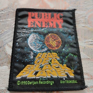 Op de afbeelding: Een rechthoekige zwarte stoffen patch met de rode tekst PUBLIC ENEMY bovenaan. Daaronder toont een afbeelding een witte maan en een oranje planeet tegen een donkere achtergrond. De gele tekst FEAR OF A BLACK PLANET staat onderaan.