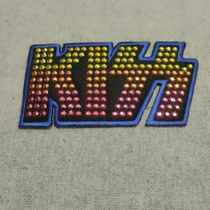 May include: A black patch featuring the word "KISS" in a stylized font. The letters are outlined in blue and filled with rhinestones, shifting from pink to yellow. This patch is designed for application on clothing or accessories.