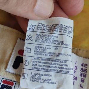 Puede incluir: Primer plano de una etiqueta de ropa con instrucciones de lavado y el logotipo de Fila. La etiqueta incluye s&iacute;mbolos para lavar a m&aacute;quina, no usar lej&iacute;a, planchar a baja temperatura y limpieza en seco. Tambi&eacute;n se muestra informaci&oacute;n sobre tallas.