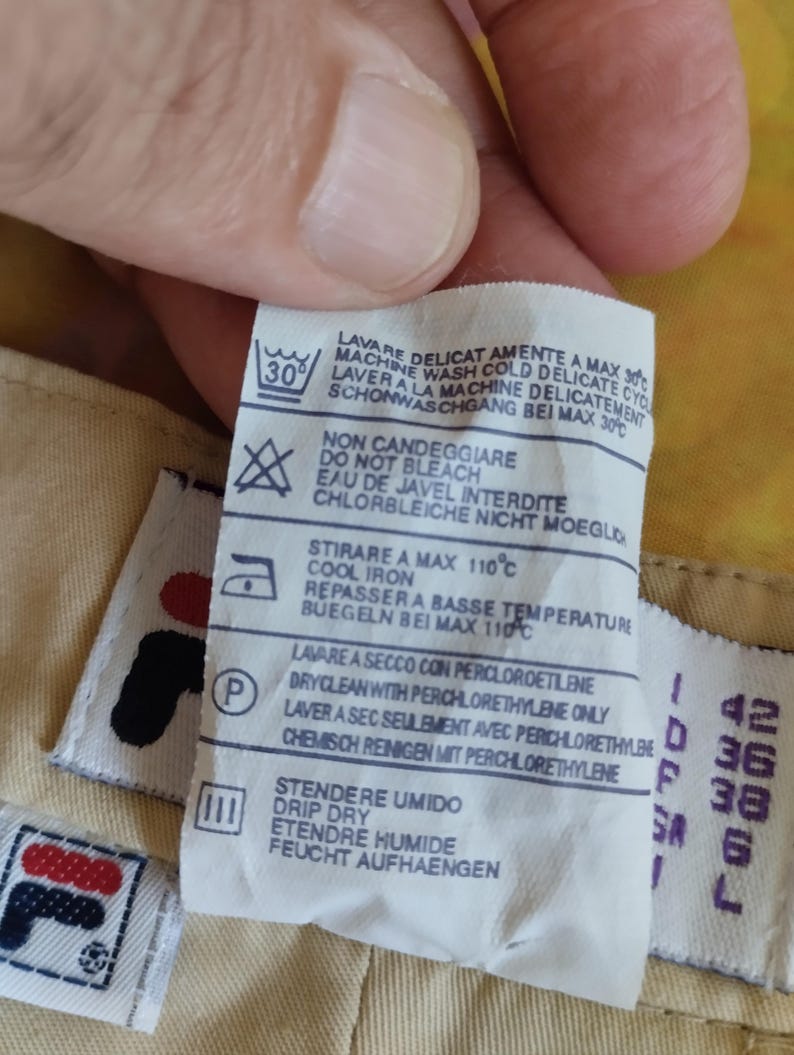 Puede incluir: Primer plano de una etiqueta de ropa con instrucciones de lavado y el logotipo de Fila. La etiqueta incluye s&iacute;mbolos para lavar a m&aacute;quina, no usar lej&iacute;a, planchar a baja temperatura y limpieza en seco. Tambi&eacute;n se muestra informaci&oacute;n sobre tallas.