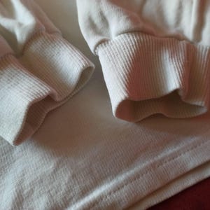 May include: Close-up of a cream-coloured long-sleeved shirt. The image highlights the ribbed cuffs and the fabric's texture. The cuffs are folded over, and the shirt's body is visible below.
