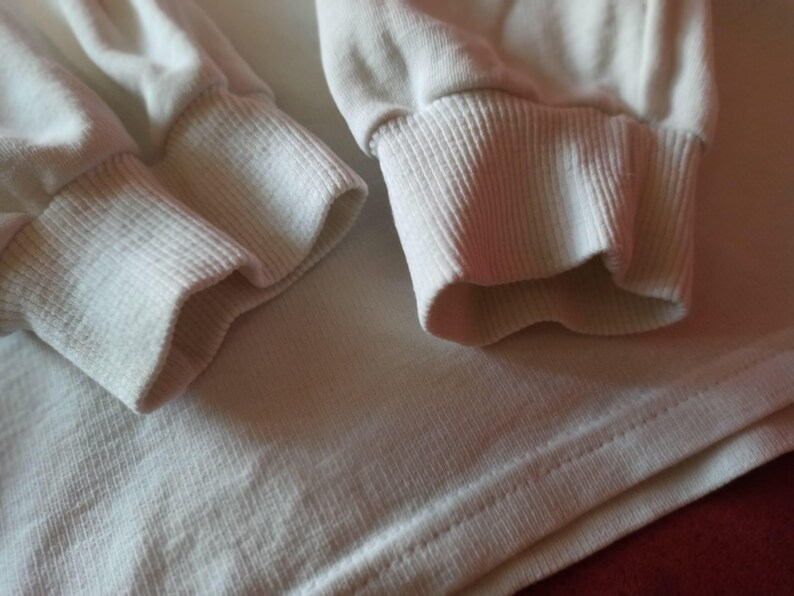 May include: Close-up of a cream-coloured long-sleeved shirt. The image highlights the ribbed cuffs and the fabric's texture. The cuffs are folded over, and the shirt's body is visible below.