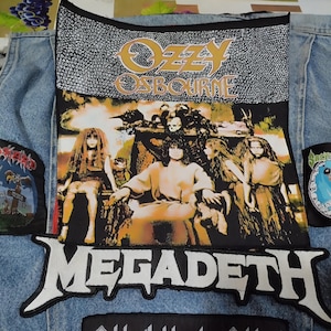 Op de afbeelding: Een lichtblauw spijkergilet met een grote rechthoekige Ozzy Osbourne-patch met een afbeelding van figuren, een witte Megadeth-logopatch, een Tankard-patch aan de linkerkant en een Sacred Reich Nicaragua-patch aan de rechterkant van het kledingstuk.