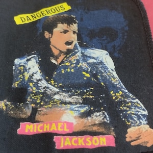 May include: A rectangular fabric patch with a black border featuring a graphic of Michael Jackson in a blue jacket with yellow accents. The text DANGEROUS appears in a yellow banner at the top, while MICHAEL JACKSON is printed in pink at the bottom.