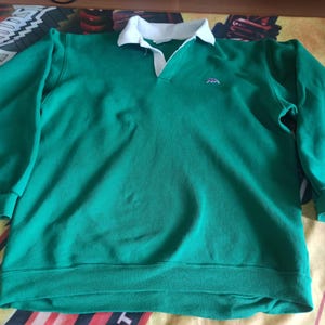 Op de afbeelding: Groen sweatshirt met een witte kraag en een klein geborduurd logo. De top met lange mouwen heeft een V-hals en geribde manchetten en zoom. De stof lijkt een zacht, comfortabel materiaal te zijn, geschikt voor vrijetijdskleding.