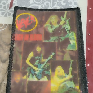 Puede incluir: Un parche tejido negro y rojo con el nombre de la banda "Slayer" y el título del álbum "Reign in Blood" en texto blanco. El parche presenta un collage de imágenes de los miembros de la banda actuando.