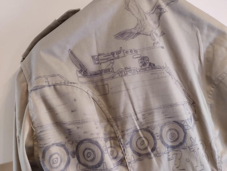 May include: A beige jacket with a drawing of a military vehicle on the back. The vehicle has a cannon on top and wheels with treads. The drawing is in black ink and includes the text "Model 127".
