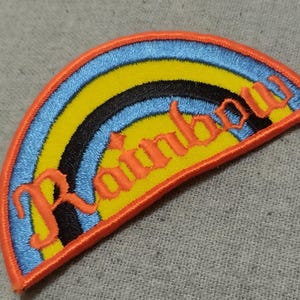 Może przedstawiać: Haftowana naszywka z tęczą z niebieskimi, żółtymi i czarnymi paskami. Słowo "Rainbow" jest wyszyte na pomarańczowo. Naszywka ma pomarańczową obwódkę i jest umieszczona na szarym tle z tkaniny.