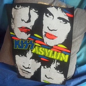 Pode incluir: Uma t-shirt cinzenta com uma impressão gráfica a preto e branco dos quatro membros da banda KISS. O design inclui o texto KISS ASYLUM em letras azuis e amarelas, acentuado com detalhes de pinceladas horizontais vermelhas, azuis e verdes.