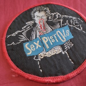 May include: Round black patch with a red border featuring a stylized illustration of a person with blood on their face. The words "Sex Pistols" are embroidered on a blue banner across the image.