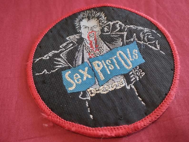 May include: Round black patch with a red border featuring a stylized illustration of a person with blood on their face. The words "Sex Pistols" are embroidered on a blue banner across the image.