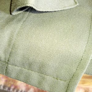 May include: Close-up of a green fabric jacket with a button closure. The fabric has a textured weave and the button is a dark brown color.