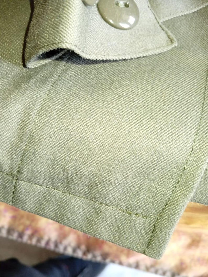 May include: Close-up of a green fabric jacket with a button closure. The fabric has a textured weave and the button is a dark brown color.