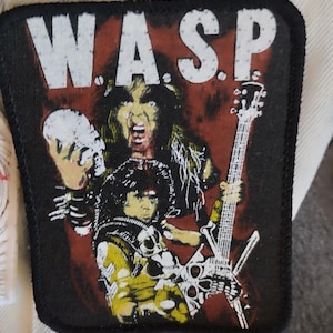 May include: A black and white embroidered patch with the word "W.A.S.P." in white lettering. The patch features a black and white image of a rock band member holding a guitar and a skull.