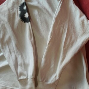 May include: White long-sleeved shirt featuring a black number 8 on the left sleeve. The shirt has ribbed cuffs at the wrists and a crew neck. The fabric appears to be a soft, lightweight material.