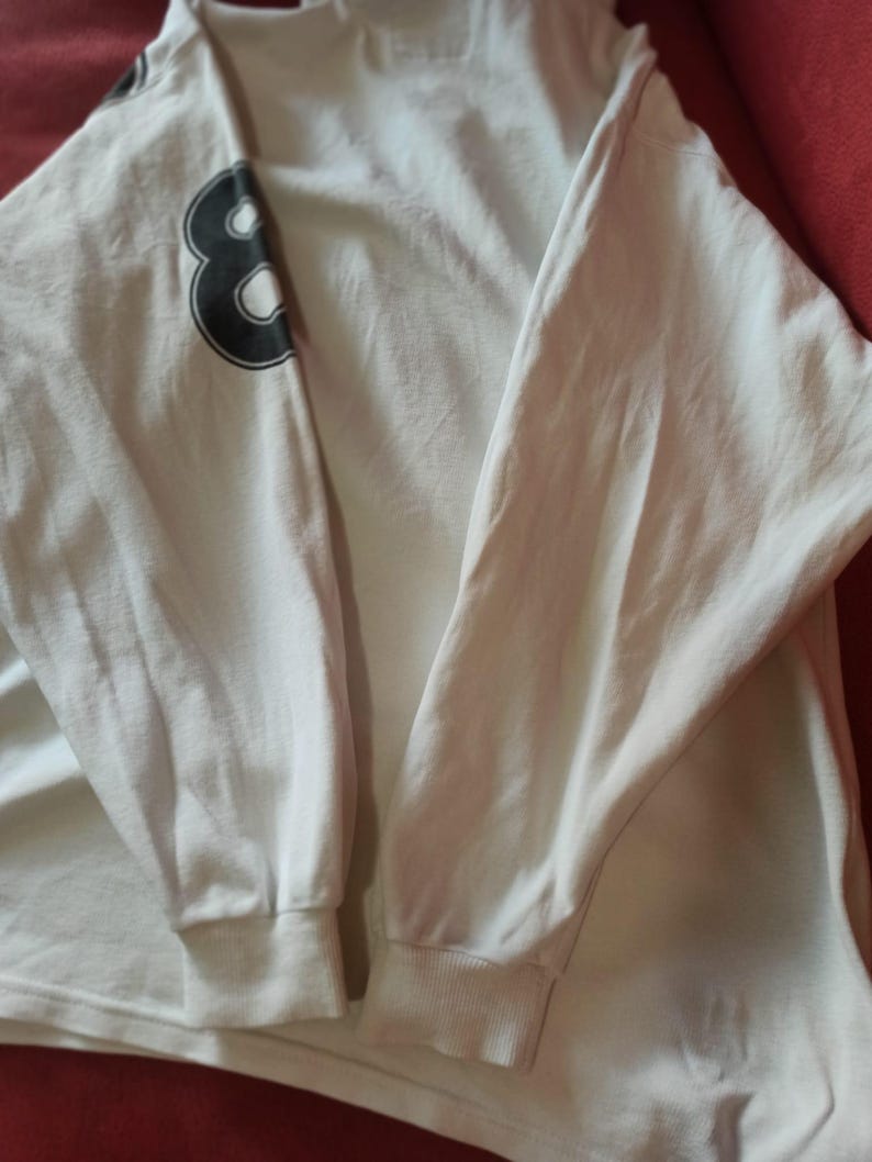 May include: White long-sleeved shirt featuring a black number 8 on the left sleeve. The shirt has ribbed cuffs at the wrists and a crew neck. The fabric appears to be a soft, lightweight material.
