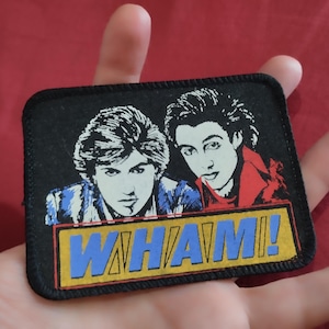 Pode incluir: Um emblema retangular bordado com uma borda preta apresentando uma ilustração gráfica de duas pessoas com cabelos escuros. Abaixo das figuras, uma faixa retangular amarela contém o texto azul WHAM! em uma fonte negrito e estilizada.