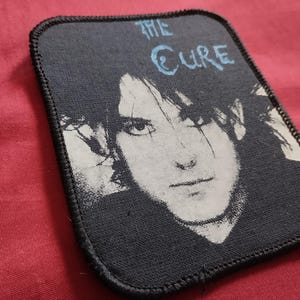 Pode incluir: Patch quadrado com um retrato a preto e branco de uma pessoa, com o texto "THE CURE" em azul claro. O patch tem uma borda preta e é provavelmente feito de tecido. O fundo é de cor vermelha sólida.