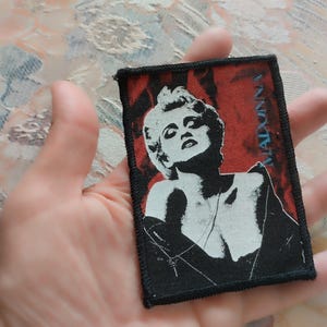 Może przedstawiać: Prostokątna naszywka z tkaniny z czarno-białą grafiką przedstawiającą Madonnę na głębokim czerwonym tle. Nazwa MADONNA jest nadrukowana pionowo na niebiesko po prawej stronie. Naszywka posiada grubą, czarną, obszytą krawędź.