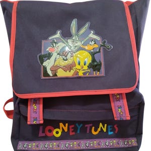 May include: A dark purple backpack featuring Looney Tunes characters Bugs Bunny, Daffy Duck, Tweety Bird, and others. The backpack has red trim, adjustable straps, and the words "LOONEY TUNES" in colorful letters. A decorative band with cartoon characters is at the bottom.