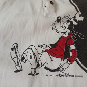 May include: White polo shirt with a cartoon image of Goofy wearing a red jacket and white trousers. The graphic is outlined in black. The text "&copy; 87 The Walt Disney Company" is printed below the illustration.