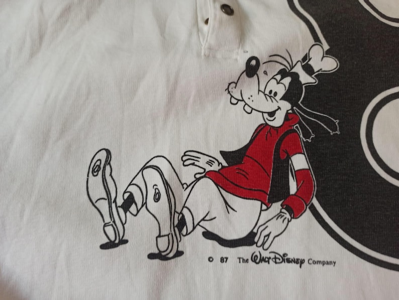 May include: White polo shirt with a cartoon image of Goofy wearing a red jacket and white trousers. The graphic is outlined in black. The text "&copy; 87 The Walt Disney Company" is printed below the illustration.