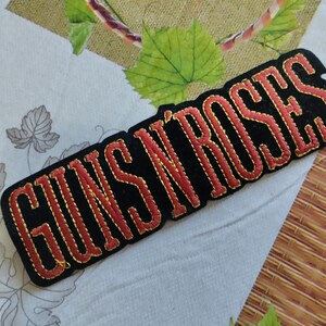 May include: A black embroidered patch featuring the words "GUNS N' ROSES" in red, with a gold outline. The patch has a rectangular shape with rounded edges. It rests on patterned fabric.