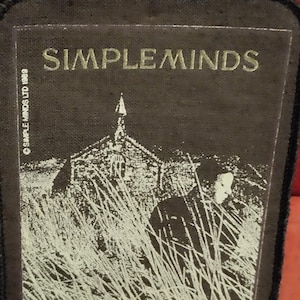 以下が含まれることがあります： 金色の文字で「SIMPLEMINDS」というバンド名が書かれた白黒のパッチ。パッチには教会と長い草の中にいる人物が描かれています。「© SIMPLE MINDS LTD 1989」の文字も表示されています。