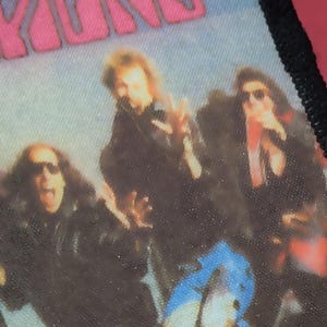 Op de afbeelding: Een rechthoekige stoffen patch met een zwarte gestikte rand met een gedrukte afbeelding van een rockband. De afbeelding toont figuren in donkere kleding tegen een lichte achtergrond met de roze tekst ARRIONS zichtbaar aan de bovenrand.