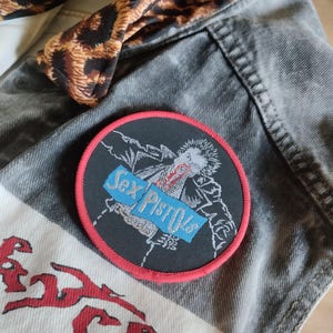May include: A black and red Sex Pistols patch is on a gray denim jacket. The patch features a graphic of a person and the band's name in blue text. A leopard print fabric is visible.