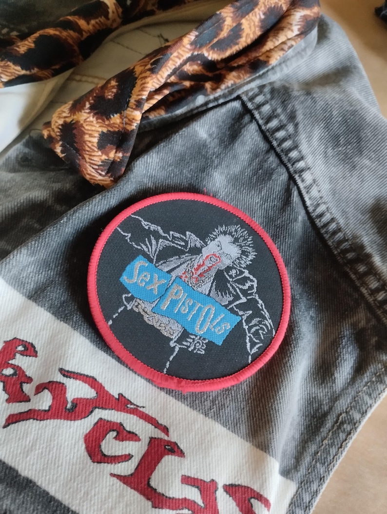 May include: A black and red Sex Pistols patch is on a gray denim jacket. The patch features a graphic of a person and the band's name in blue text. A leopard print fabric is visible.