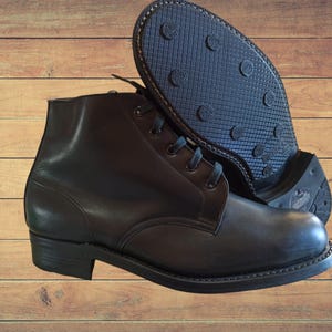 Puede incluir: Botines de cuero negro con cordones. Las botas tienen una suela resistente y un tacón bajo. Una bota está de pie, mientras que la otra muestra el dibujo de la suela. El fondo es de madera.