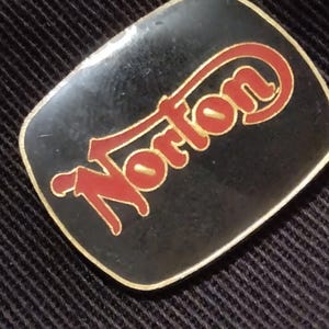 May include: A black and gold metal pin with the word "Norton" in red lettering.