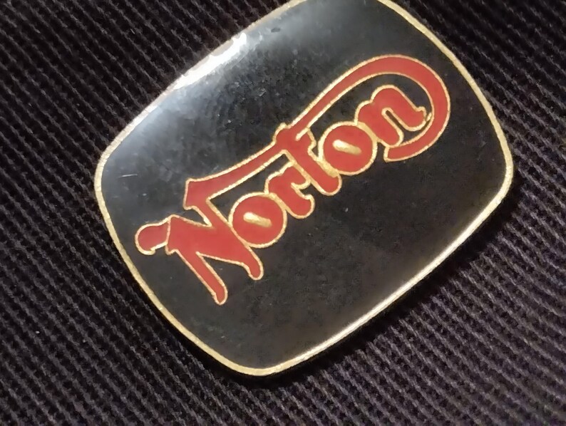May include: A black and gold metal pin with the word "Norton" in red lettering.