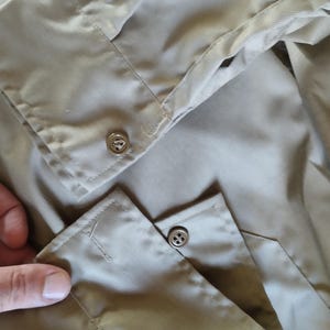 May include: Close-up of the cuff of a beige long-sleeved shirt with two buttons. The buttons are metal and have a four-hole design.