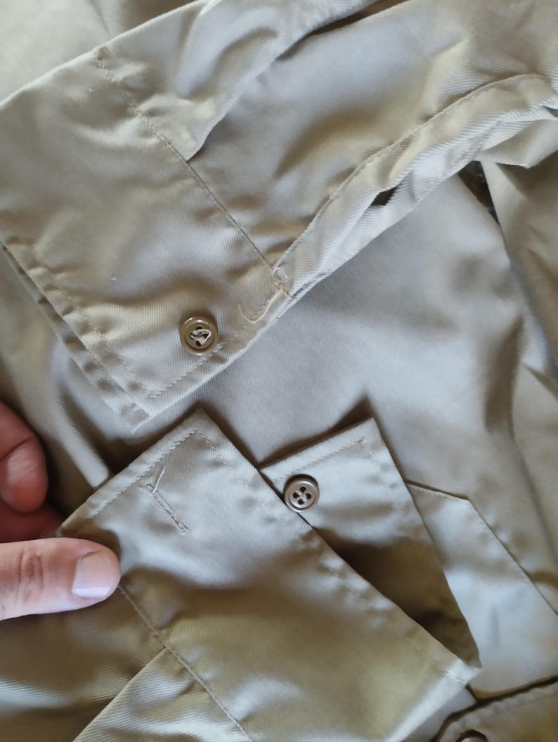 May include: Close-up of the cuff of a beige long-sleeved shirt with two buttons. The buttons are metal and have a four-hole design.
