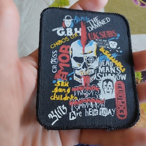 May include: A black, rectangular patch with a skull graphic and text in white, red, and yellow. The patch includes band names such as "The Damned" and "UK Subs". The design has a punk rock aesthetic.