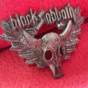 May include: A silver metal emblem with the words "Black Sabbath" above a skull with horns and wings. The emblem has a distressed finish and is set against a red background, giving it a vintage look.