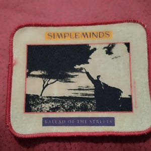 May include: A rectangular fabric patch with rounded corners and a red stitched border. The centre features a black and white graphic of a person reaching towards a tree. The text SIMPLE MINDS appears at the top and BALLAD OF THE STREETS at the bottom.