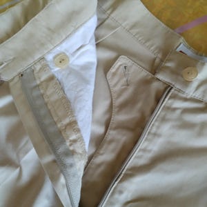 Puede incluir: Primer plano de pantalones color caqui con cremallera abierta y cierre de bot&oacute;n. Los pantalones tienen un dise&ntilde;o cl&aacute;sico con un forro blanco visible y una cintura abotonada. La tela parece ser una mezcla de algod&oacute;n duradera.