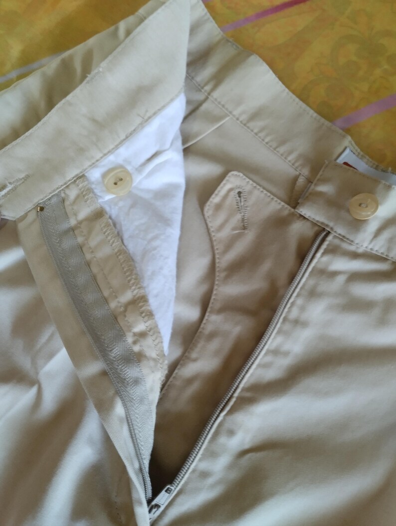 Puede incluir: Primer plano de pantalones color caqui con cremallera abierta y cierre de bot&oacute;n. Los pantalones tienen un dise&ntilde;o cl&aacute;sico con un forro blanco visible y una cintura abotonada. La tela parece ser una mezcla de algod&oacute;n duradera.