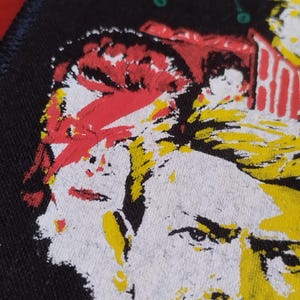 Op de afbeelding: Close-up van een zwart embleem met een portret in pop-artstijl in geel, rood en wit. De afbeelding lijkt van een persoon te zijn. De patch heeft een donkerblauwe rand.