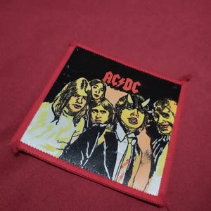 May include: Square patch featuring the band AC/DC. The patch has a black background with a group photo of the band members. The band's name is in red letters at the top. The patch has a red border.