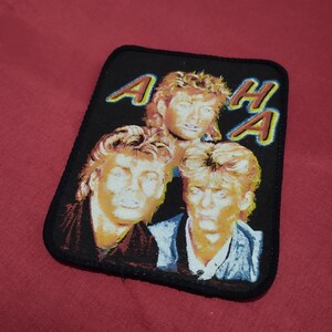 Könnte beinhalten: Schwarzer rechteckiger Aufnäher mit der Band A-ha. Der Aufnäher hat einen schwarzen Rand und den Namen der Band in Gelb und Blau. Der Aufnäher zeigt drei Bandmitglieder mit blonden Haaren.