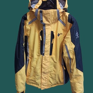 Peut inclure: Veste à capuche jaune et bleu marine avec fermeture éclair. La veste présente des détails argentés réfléchissants, un logo sur la poitrine et un logo sur la manche. Elle est suspendue à un cintre noir.