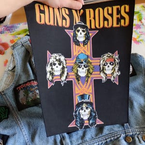 Può includere: Una toppa in tessuto nero con il logo Guns N' Roses in testo giallo e arancione sopra un design a croce viola. La croce mostra cinque teschi illustrati con capelli lunghi e vari cappelli, posizionata sopra un gilet di jeans azzurro.