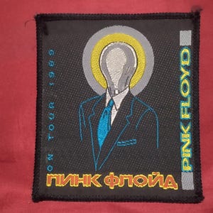 Könnte beinhalten: Ein rechteckiger, schwarzer Stickaufnäher mit einer glühbirnenköpfigen Figur im Anzug mit blauer Krawatte und einem gelb-grauen Heiligenschein. Der Text lautet PINK FLOYD, ON TOUR 1989, ergänzt durch kyrillische Schriftzeichen in Gelb und Rot.
