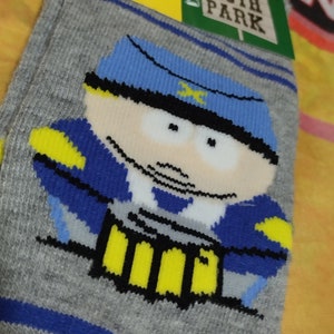 以下が含まれることがあります： 青い帽子に黄色の星をつけた青と黄色の漫画のキャラクターが描かれた灰色の靴下。キャラクターはテレビ番組サウスパークのものです。靴下にはコメディセントラルのロゴと「サウスパーク」の文字がタグに書かれています。